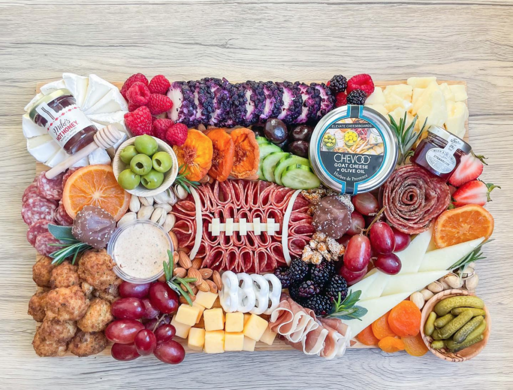 Football Game Day grazing board with cold cuts in the shape of a football, dips, veggies, hot honey, goat cheese and fruit
