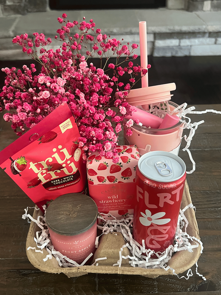 A pink themed gift basket with a pink drink, pink candy, pink candle and pink flowers.